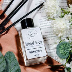 Peut inclure: Un flacon diffuseur d'ambiance en verre transparent avec un bouchon noir, étiqueté "Midnight Amber" avec des senteurs de jasmin, ambre et bois dorés. L'étiquette indique également "Room Diffuser" et "150g | 7oz". L'image comprend des bâtonnets noirs, des fleurs blanches et des feuilles vertes.