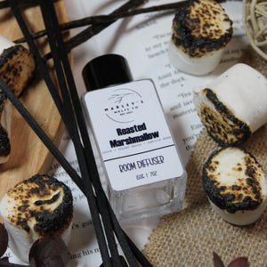 Może przedstawiać: Dyfuzor do pomieszczeń "Roasted Marshmallow" z czarnymi patyczkami, otoczony prażonymi piankami i strąkami wanilii. Dyfuzor ma pojemność 50 ml.