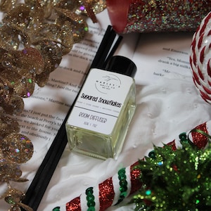 Może przedstawiać: Świąteczna martwa natura z czerwoną świecą z brokatem, dyfuzorem do pomieszczeń z napisem "Sugared Snowflakes" i elementami dekoracyjnymi. Dyfuzor znajduje się w przezroczystej szklanej butelce. Scena zawiera laskę cukrową i złote akcenty brokatu.