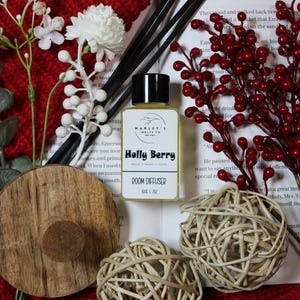Könnte beinhalten: Eine Raumduftflasche mit schwarzem Deckel und dem Aufdruck "Holly Berry". Die Flasche ist von dekorativen Elementen umgeben, darunter rote Beeren, weiße Blüten und geflochtene Kugeln, alles vor einem roten Strickhintergrund.