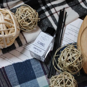 Może przedstawiać: Martwa natura z dyfuzorem do pomieszczeń "Flannel", patyczkami trzcinowymi i dekoracyjnymi kulami z plecionki. Elementy spoczywają na tkaninie w kratę w odcieniach czerni, bieli i błękitu. Obecna jest również drewniana taca i zadrukowany papier.