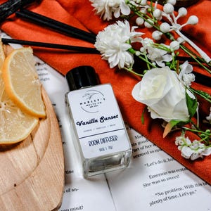 Może przedstawiać: Przezroczysta szklana butelka dyfuzora do pomieszczeń z czarną nakrętką, z napisem "Vanilla Sunset". Etykieta głosi również "Room Diffuser" i "150C 1 7oz". Butelka stoi na książce obok plasterków cytryny, kwiatów i pomarańczowej tkaniny.