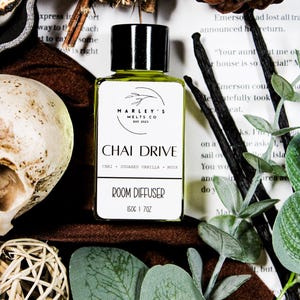 Może przedstawiać: Dyfuzor do pomieszczeń z czarną nakrętką i białą etykietą z napisem "CHAI DRIVE" z wymienionymi poniżej składnikami. Butelka otoczona jest elementami dekoracyjnymi, w tym liśćmi, laskami wanilii i czaszką.