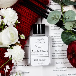 Może przedstawiać: Przezroczysta szklana butelka dyfuzora do pomieszczeń z czarną nakrętką, z etykietą "Apple Slices" od "Marley's Melts Co." Butelka otoczona jest białymi kwiatami, czerwonym dzianinowym przedmiotem i książką. Dyfuzor zawiera 30 ml płynu.