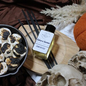 Może przedstawiać: Martwa natura o tematyce Halloween z kwadratową szklaną butelką "Doom Diffuser" z czarnymi trzcinami, drewnianą tacą i żeliwną patelnią z opieczonymi piankami. Obecne są również dekoracyjne czaszki i dynia.