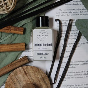 Może przedstawiać: Butelka dyfuzora do pomieszczeń z czarną nakrętką i białą etykietą z napisem "Holiday Garland" z "Chai + Evergreen" poniżej. Obraz zawiera czarne patyczki, laski wanilii, laski cynamonu i drewniany podkład.