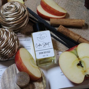 Może przedstawiać: Butelka dyfuzora do pomieszczeń z czarną nakrętką i etykietą "Cider Street". Obraz zawiera plasterki jabłka, laski cynamonu i dekoracyjne kule, sugerując jesienny motyw. Dyfuzor ma 28.35g.