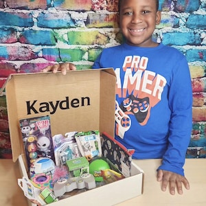 May include: A cardboard gift box with the name Kayden printed inside, containing various small toys including a Jack Skellington lollipop, a Minecraft figure, a green ball, a gray game controller, and a patterned zipper pouch.