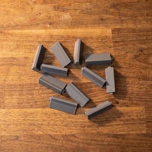 Pode incluir: Um conjunto de dez pequenos componentes retangulares de plástico cinzento. Cada peça apresenta um corpo plano e alongado com uma crista central ligeiramente elevada ao longo do comprimento e uma pequena aba saliente numa das extremidades.