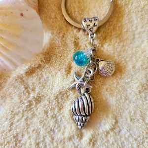 May include: A silver key chain with a heart charm and multiple sea-themed charms, including a seashell, a starfish, and a blue glass bead. The charms are hanging from a silver ring.