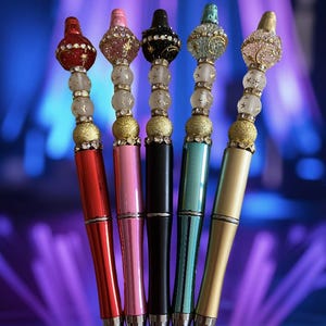 May include: Five decorative pens with jewel-encrusted tops in red, pink, black, teal, and gold. Each pen has a silver tip and a unique design with clear beads and gold accents. The pens are arranged in a fan shape.