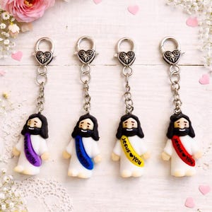 May include: Four keychains featuring a small figurine of a person with long black hair and a beard, wearing a white robe with a colored sash. The sashes are purple, blue, yellow, and red, each printed with the text Jesus loves you.