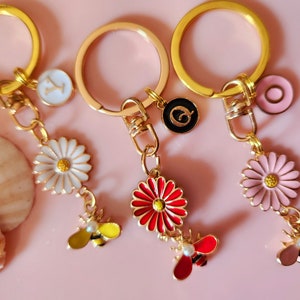 May include: Three gold keychains with charms. Each keychain has a different colored flower charm: white, red, and pink. Each flower charm has a bee charm hanging from it. The keychains also have a letter charm: Y, Q, and a pink circle.