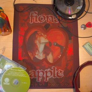 Peut inclure: Une affiche rouge et blanche avec le texte "Fiona Apple" et une pomme rouge avec un visage de femme &agrave; l'int&eacute;rieur. La femme chante dans un microphone.