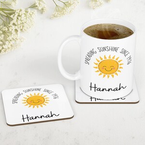 May include: A white ceramic mug filled with dark coffee and a matching square coaster. Both items feature a yellow smiling sun graphic, the text "Spreading Sunshine Since 1976" in a black sans-serif font, and the name "Hannah" in a black script font.