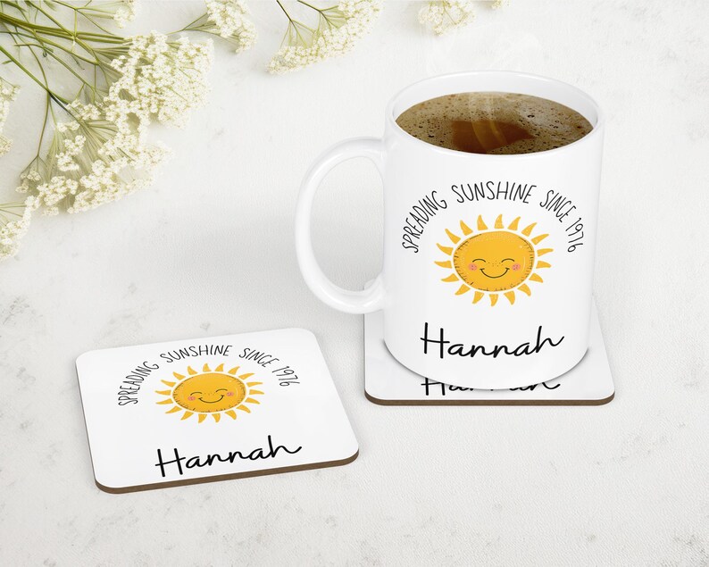 May include: A white ceramic mug filled with dark coffee and a matching square coaster. Both items feature a yellow smiling sun graphic, the text "Spreading Sunshine Since 1976" in a black sans-serif font, and the name "Hannah" in a black script font.