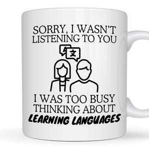 May include: A white ceramic mug featuring black text that reads, SORRY, I WASN'T LISTENING TO YOU, I WAS TOO BUSY THINKING ABOUT LEARNING LANGUAGES. Between the lines of text is a black line art icon of two people with a speech bubble containing symbols.