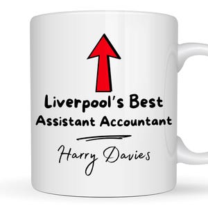May include: A white ceramic mug featuring a red upward-pointing arrow graphic above the black text "Liverpool's Best Assistant Accountant" and the name "Harry Davies" written in a cursive script font below a horizontal line.