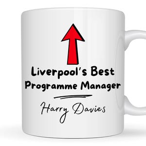 Peut inclure: Mug en céramique blanche avec une flèche rouge pointant vers le haut et le texte "Liverpool's Best Programme Manager Harry Davies" en noir. La tasse a une anse incurvée.