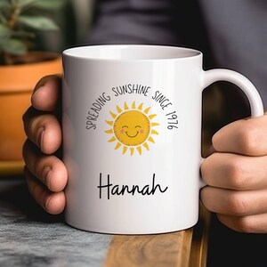 May include: A white ceramic mug featuring a yellow sun graphic with a smiling face. The text "Spreading Sunshine Since 1976" is printed in a circular arc above the sun, and the name "Hannah" is printed in black script lettering below the sun.