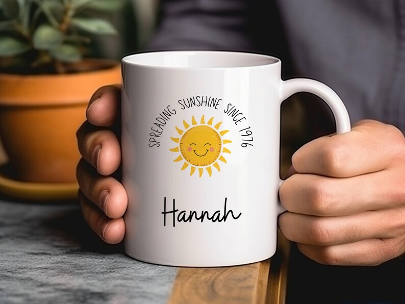 May include: A white ceramic mug featuring a yellow sun graphic with a smiling face. The text "Spreading Sunshine Since 1976" is printed in a circular arc above the sun, and the name "Hannah" is printed in black script lettering below the sun.