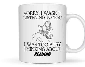 May include: A white ceramic mug featuring a black line drawing of a person sitting and reading a book. The text printed in black reads: SORRY, I WASN'T LISTENING TO YOU I WAS TOO BUSY THINKING ABOUT READING. The mug has a curved handle on the right side.