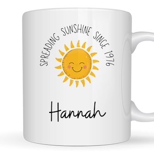 May include: A white ceramic mug featuring a yellow sun illustration with a smiling face and pink cheeks. The text reads "SPREADING SUNSHINE SINCE 1976" in a curved black font above the sun, and the name "Hannah" in black script below the sun.