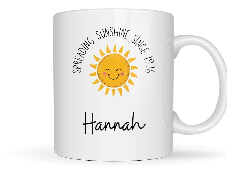 May include: A white ceramic mug featuring a yellow sun illustration with a smiling face and pink cheeks. The text reads "SPREADING SUNSHINE SINCE 1976" in a curved black font above the sun, and the name "Hannah" in black script below the sun.