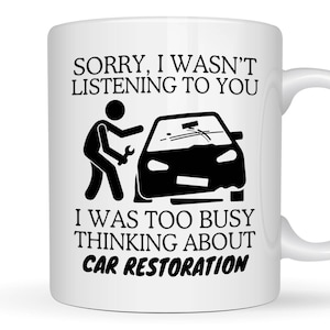 May include: A white ceramic mug featuring black text that reads SORRY, I WASN'T LISTENING TO YOU I WAS TOO BUSY THINKING ABOUT CAR RESTORATION, accompanied by a black graphic of a stick figure holding a wrench next to a car silhouette.