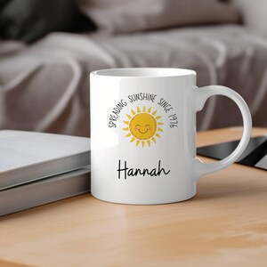 May include: A white ceramic mug featuring a yellow sun graphic with a smiling face. The text "Spreading Sunshine Since 1976" is printed in a circular arc above the sun, and the name "Hannah" is printed in black script below the sun graphic.