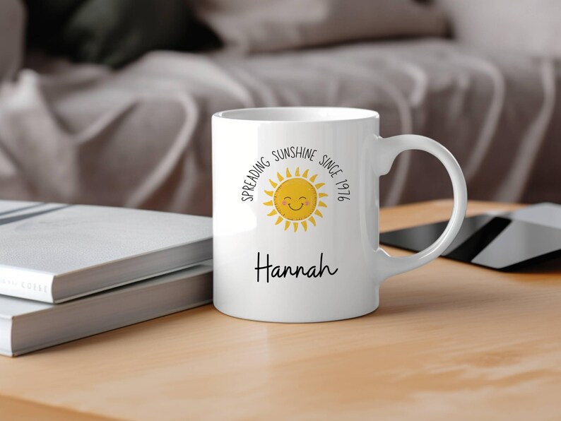 May include: A white ceramic mug featuring a yellow sun graphic with a smiling face. The text "Spreading Sunshine Since 1976" is printed in a circular arc above the sun, and the name "Hannah" is printed in black script below the sun graphic.