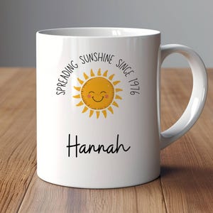 May include: A white ceramic mug featuring a yellow sun graphic with a smiling face, surrounded by the text "SPREADING SUNSHINE SINCE 1976" in black lettering. Below the sun, the name "Hannah" is printed in a black cursive script font.