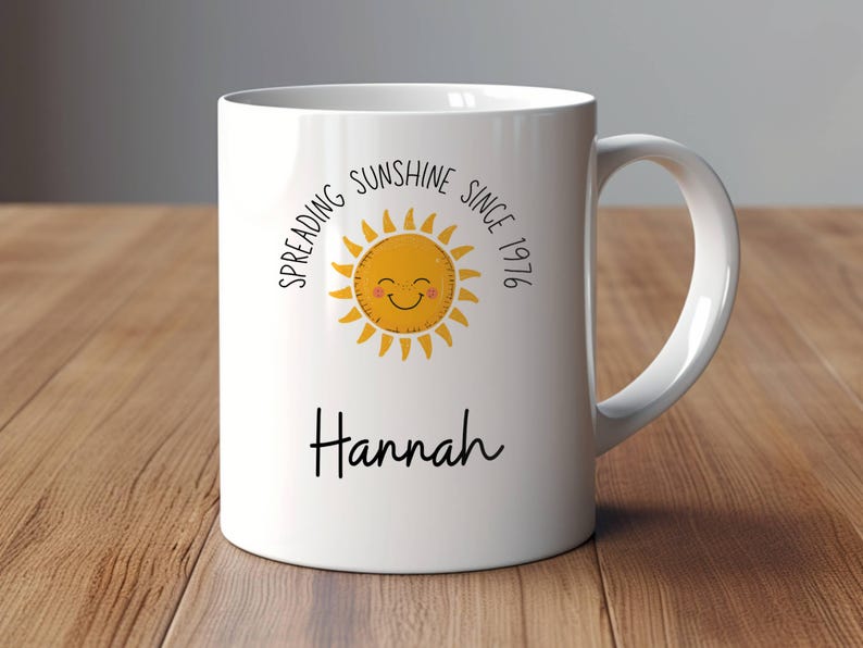 May include: A white ceramic mug featuring a yellow sun graphic with a smiling face, surrounded by the text "SPREADING SUNSHINE SINCE 1976" in black lettering. Below the sun, the name "Hannah" is printed in a black cursive script font.