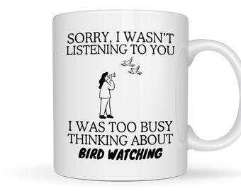 May include: A white ceramic mug featuring black text that reads, SORRY, I WASN'T LISTENING TO YOU I WAS TOO BUSY THINKING ABOUT BIRD WATCHING. The design includes a black line drawing of a person looking through binoculars at two flying birds.