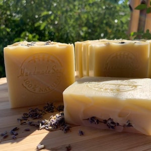 Puede incluir: Cuatro barras de jabón hecho a mano con aroma a lavanda. El jabón es de color amarillo claro y tiene un sello que dice "Handmade Soap 100% Natural". El jabón está sobre una superficie de madera con flores de lavanda secas.