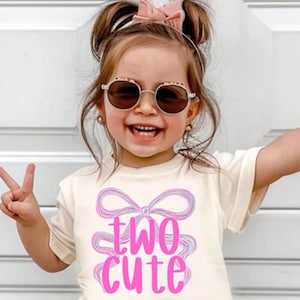 May include: A child's cream-coloured t-shirt with the pink text "two cute" and a bow design. The child is wearing denim shorts and sunglasses. The child is smiling and making a peace sign with their fingers.