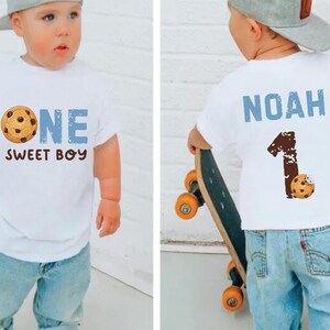 May include: A white t-shirt with the text "ONE SWEET BOY" and a cookie graphic. The child is wearing light blue jeans and a gray baseball cap. The back of the shirt has the name "NOAH" and the number "1" with a cookie graphic.