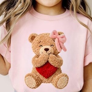 May include: A light pink t-shirt featuring a smiling teddy bear holding a red heart. The teddy bear has a pink bow and is made of a textured, plush material. The shirt has short sleeves.