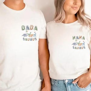 以下が含まれることがあります： お揃いの恐竜グラフィックが入ったオフホワイトのTシャツ2枚。「DADA SAURUS」と「MAMA SAURUS」の文字が遊び心のあるフォントでプリントされています。Tシャツは半袖で、綿でできているようです。