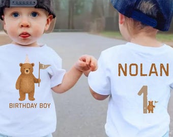 May include: Two white t-shirts with a brown bear graphic. One shirt says "BIRTHDAY BOY" and the other says "NOLAN 1". The bear is holding a flag. The shirts are worn by a child.