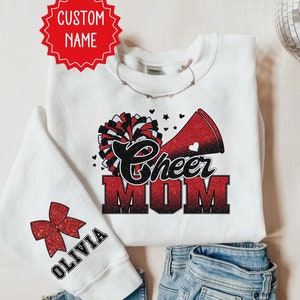 Könnte beinhalten: Weißer Pullover mit einem roten und schwarzen Glitzerdesign, auf dem "Cheer Mom" mit einer roten Schleife und einem Megaphon steht. Der Ärmel hat eine rote Schleife mit dem Namen "Olivia" darauf.