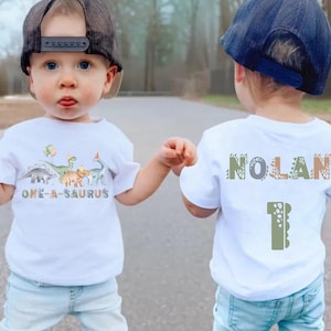 May include: White t-shirt with a dinosaur theme. The front of the shirt says "ONE-A-SAURUS" with dinosaur graphics. The back of the shirt says "NOLAN" and has the number "1". The shirt is worn by a child.