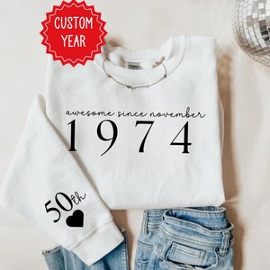 May include: White sweatshirt with black text that reads "awesome since november 1974". The sleeve has a black heart with the text "50th" above it.