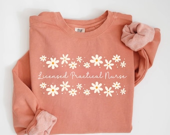 May include: Terracotta-colored sweatshirt with white daisy floral design and the text "Licensed Practical Nurse". The sweatshirt has a crew neck and long sleeves. A matching scrunchie is placed on the right side of the sweatshirt.