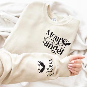 Könnte beinhalten: Ein cremefarbener Pullover mit dem Text "Mom of an angel" und einem kleinen Engelsflügel-Design. Der Ärmel hat ein kleines Engelsflügel-Design und den Namen "Olivia" darunter.
