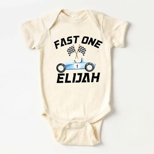 May include: A cream-colored baby onesie with a blue race car and checkered flags. The text "FAST ONE" is above the car and the name "ELIJAH" is below the car.