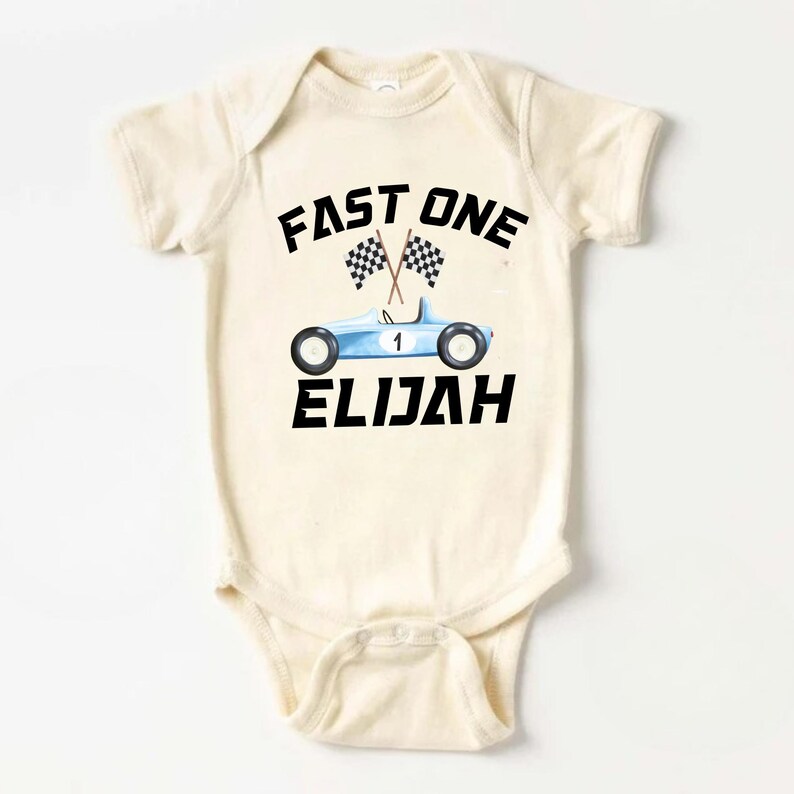 May include: A cream-colored baby onesie with a blue race car and checkered flags. The text "FAST ONE" is above the car and the name "ELIJAH" is below the car.