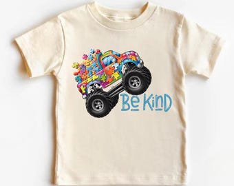 May include: A light beige t-shirt with a colorful monster truck made of puzzle pieces. The truck is driving up a hill and the words "Be Kind" are printed in blue below the truck.