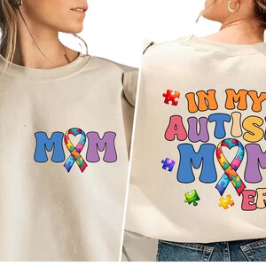 May include: A beige sweatshirt with a colorful puzzle piece ribbon and the text "MOM" on the front. The back of the sweatshirt has the text "IN MY AUTISM MOM ERA" with a colorful puzzle piece ribbon.
