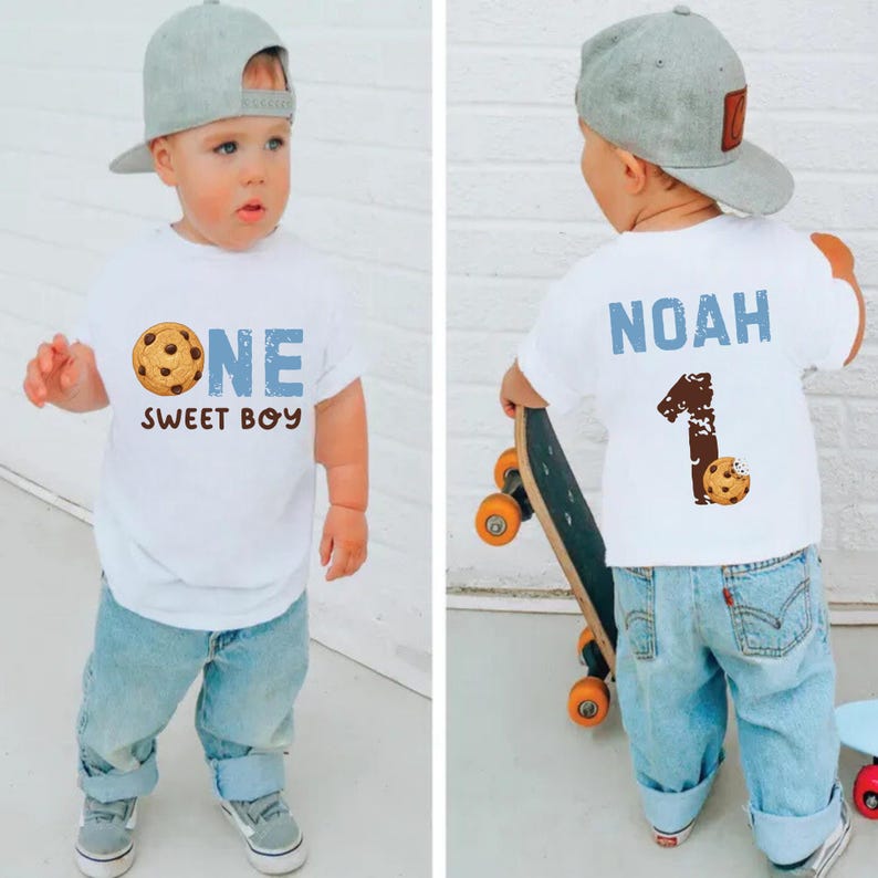 May include: A white t-shirt with the text "ONE SWEET BOY" and a cookie graphic. The child is wearing light blue jeans and a gray baseball cap. The back of the shirt has the name "NOAH" and the number "1" with a cookie graphic.